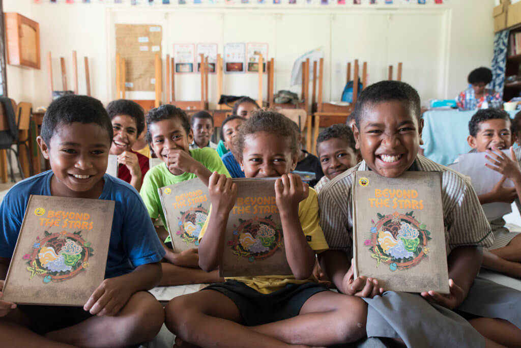 A class of smiling Fijian children hold up the Beyond the Stars storybook at school