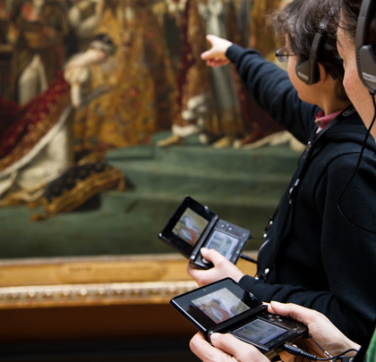 Two people hold Nintendo devices connected to headphones as they learn about an artwork from the Lourve